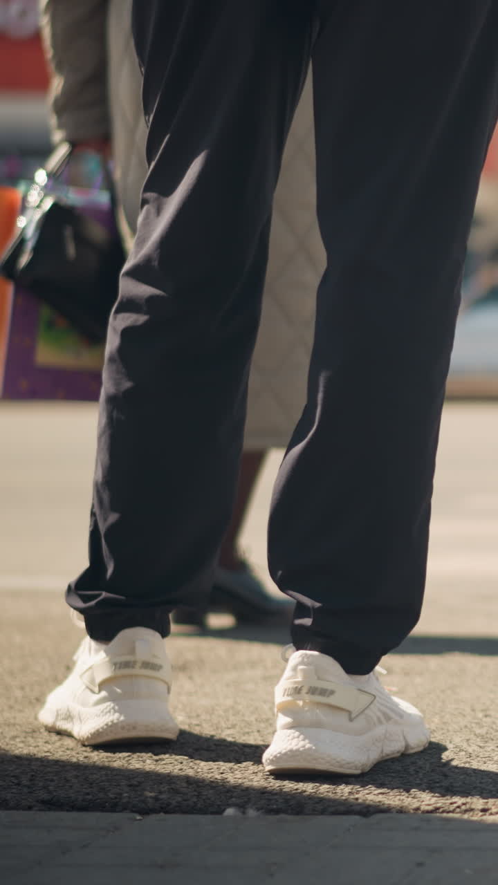 Uomo bianco in piedi su un marciapiede illuminato dal sole, primo piano dal basso angolo di gambe e sneakers bianche, pantaloni neri risvoltati, superficie in cemento ruvida, leggera ombra proiettata, posa statica che suggerisce attesa