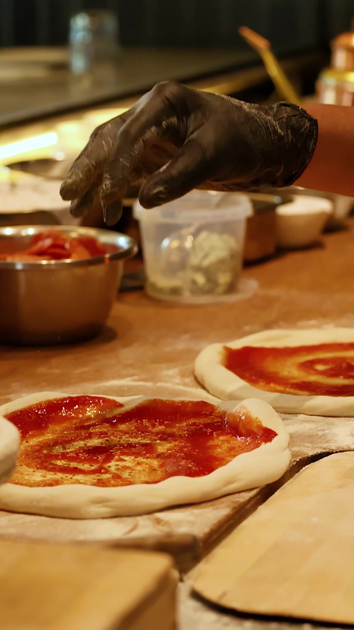 Primo piano di mani guantate che condiscono l'impasto crudo della pizza con salsa di pomodoro in un ristorante.