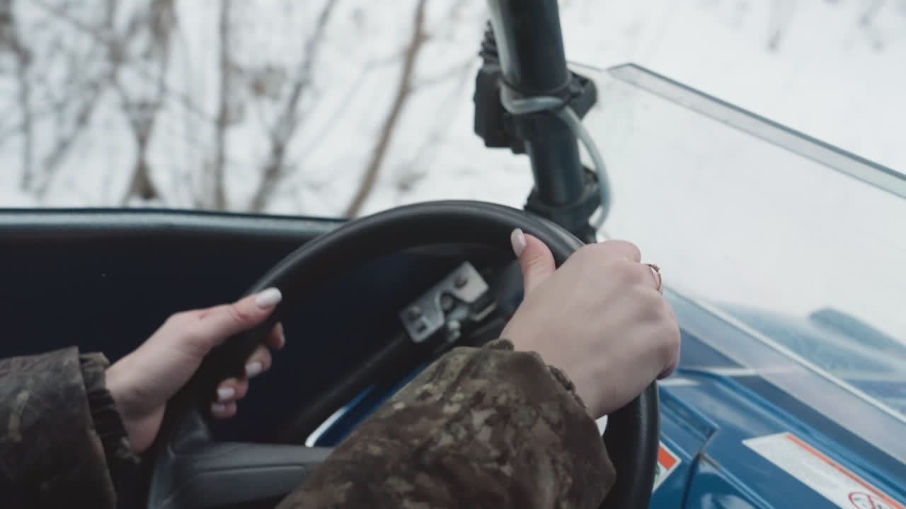 Mãos em close-up no volante de um quadriciclo, controle da trilha de inverno com jaqueta camuflada, painel azul e manopla texturizada, curvas precisas na neve, concentração focada do operador e movimento sutil.