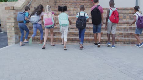 diverse group of schoolchildren wearing backpacks smiling and running upstairs up to school