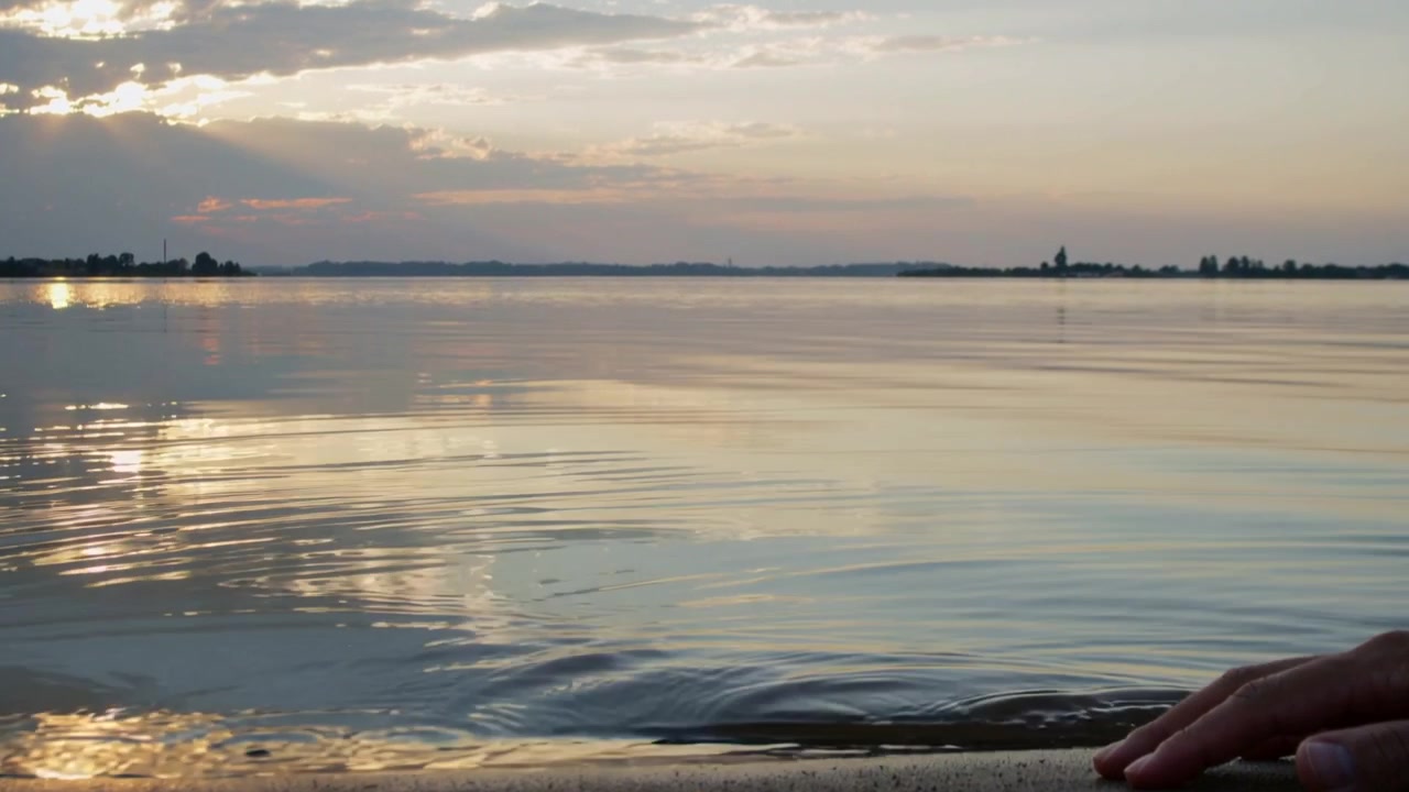 Una mano destra che tocca l'acqua creando increspature sulla riva sabbiosa al tramonto, con il riflesso del sole.