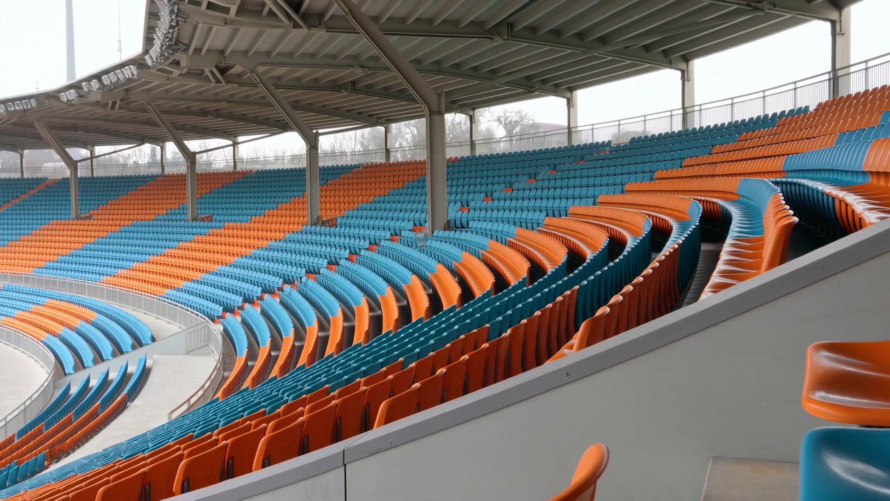 Wide view of empty covered stadium stands featuring alternating orange and teal plastic seats arranged in curved tiers and aisles; metal roof structure