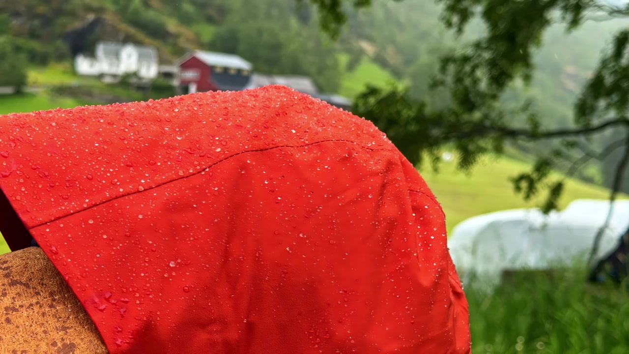 Person i orange regnjakke står i regnfuldt landskab med landbrugsbygninger
