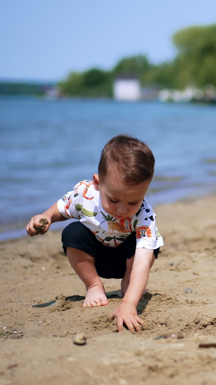 Barefoot Caucasian baby plays peacefully on the river bank. Little boy is interested in wet sand. Blurred backdrop. Vertical video