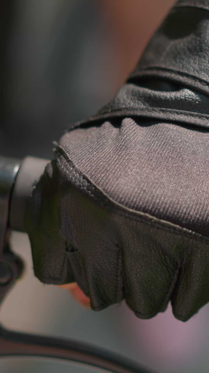 장갑을 입은 손으로 자전거 손잡이를 잡고 브레이크 레버를 들고 있는 개인의 클로즈업, 가죽 장갑에서 빛이 미묘하게 반사되고, 배경에는 녹색의 부드러운 흐릿함이 나타납니다.