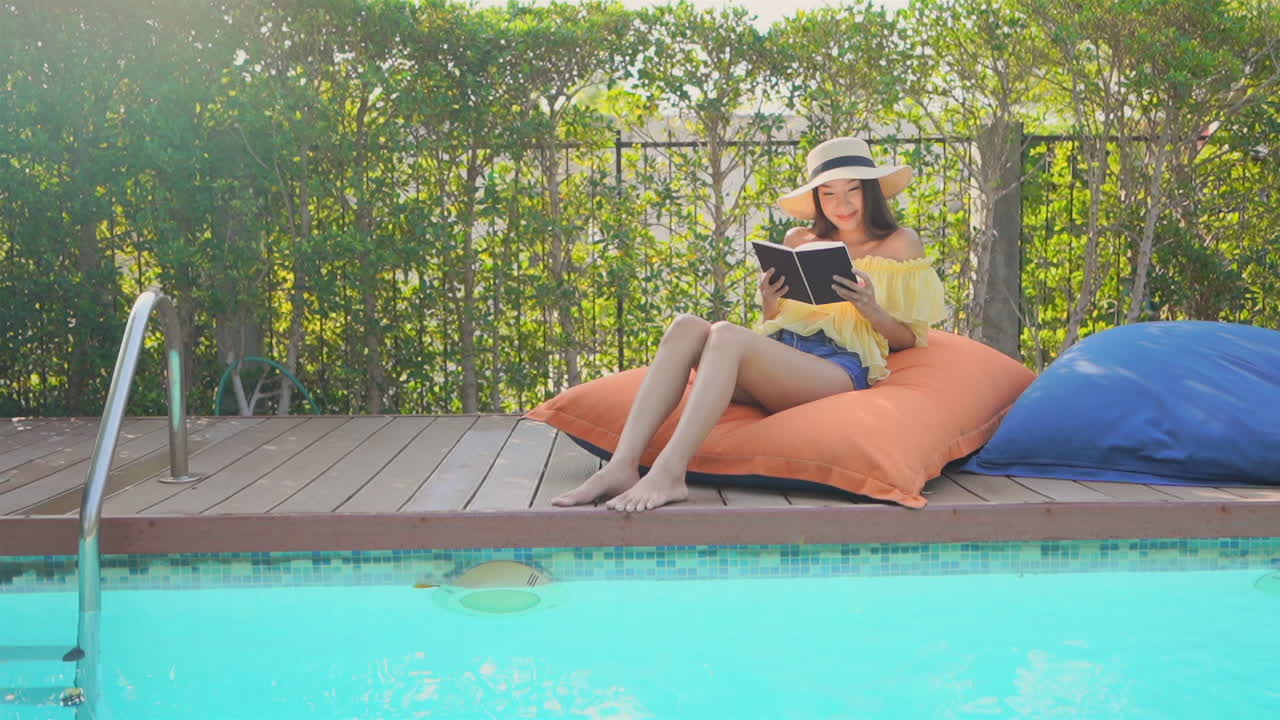Asian woman reading on a cushion beside a tropical hotel pool