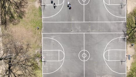 An-aerial-view-over-4-people-playing-basketball-on-a-court-surrounded-by-dry-trees-on-a-sunny-day