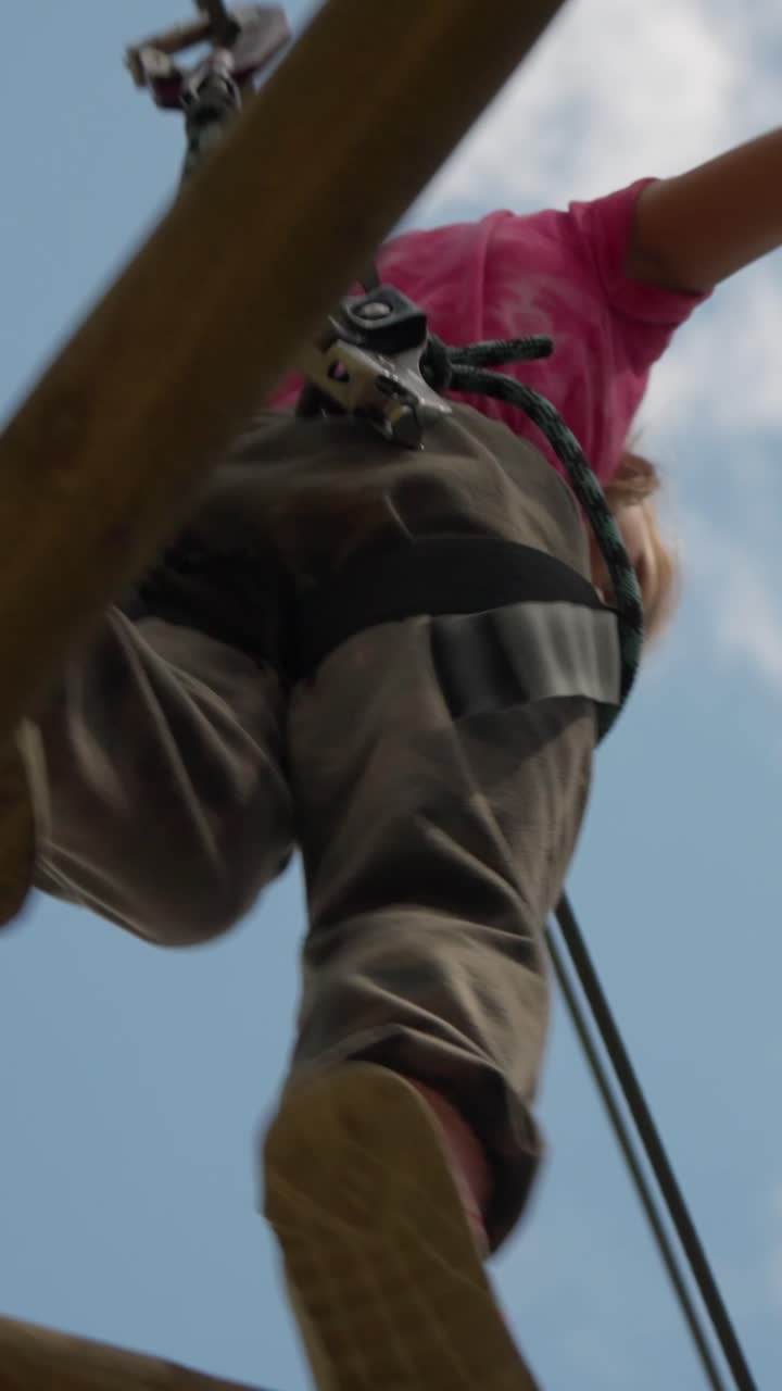Person engaging in an adventurous outdoor activity, possibly ziplining or on a rope course, seen from a low angle against a clear blue sky. Focus on feet and safety harness highlights thrilling rec