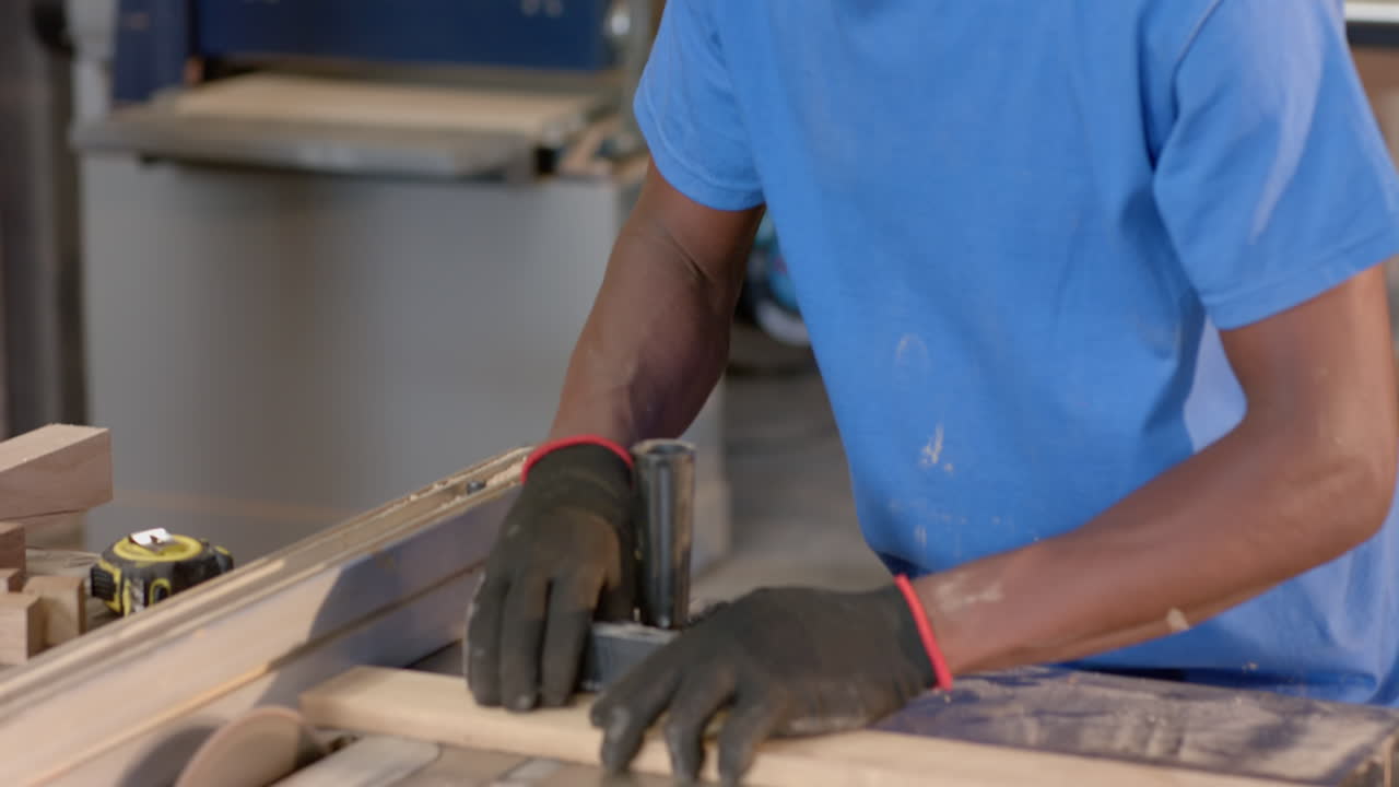 African American man wearing gloves pushing plank through table saw at workshop, with push block
