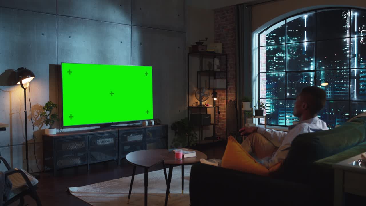 흑인 남성이 집에서 라운지 소파에 앉아서 휴식을 취하면서 녹색 화면으로 tv를보고있는 저녁 촬영. 스마트 tv를보고 현대 리모컨을 사용하는 남자