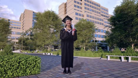 full body of asian woman student graduates in cap and gown smiling and pointing to diploma in her hand while standing in front of a magnificent university building