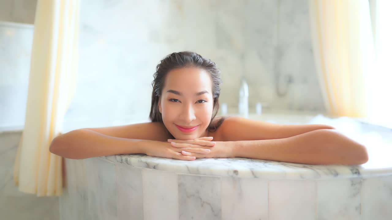 Woman smiling and resting chin on hands looking out of large bathtub filled with warm water and soap suds. Lady smiles at camera while soaking in marble tub of luxury resort suite