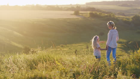 Ein-Kleines-Cooles-Mädchen-Mit-Blonden-Haaren-Zeigt-Mama-Einen-Wunderschönen-Sonnenuntergang-über-Grünen-Feldern-Rückansicht