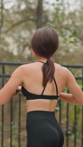 Lady performing fitness exercise outdoors in front of iron fence with trees in background, she is stretching her arms while maintaining a healthy and active lifestyle in natural environment