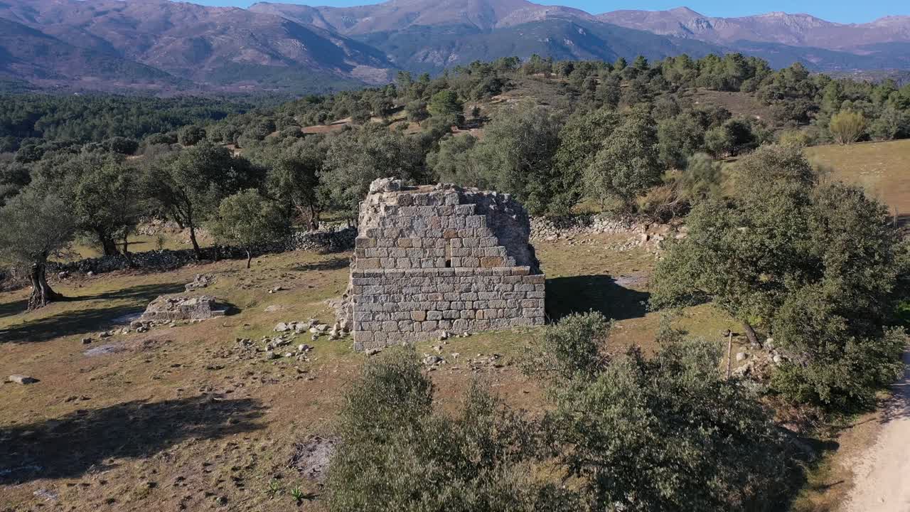 Volo ascendente con un drone che visualizza una torre medievale in rovina in una fattoria con muri in pietra accanto a una strada sterrata con uno sfondo di montagne in inverno ad Avila, Spagna