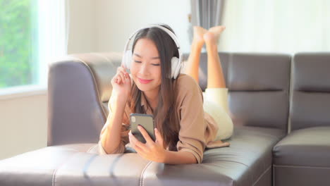 a young woman playfully kicks her feet while lying on the couch as she listens to music through her smartphone and headphones