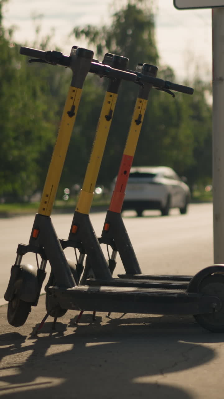 햇볕이 잘 드는 연석에 주차된 노란색 스쿠터, 교외 도로 옆에 세워진 세 대의 도크리스 전동 스쿠터와 지나가는 자동차, 나무가 늘어선 배경, 긴 그림자와 따뜻한 늦은 오후 햇살이 느껴져요.