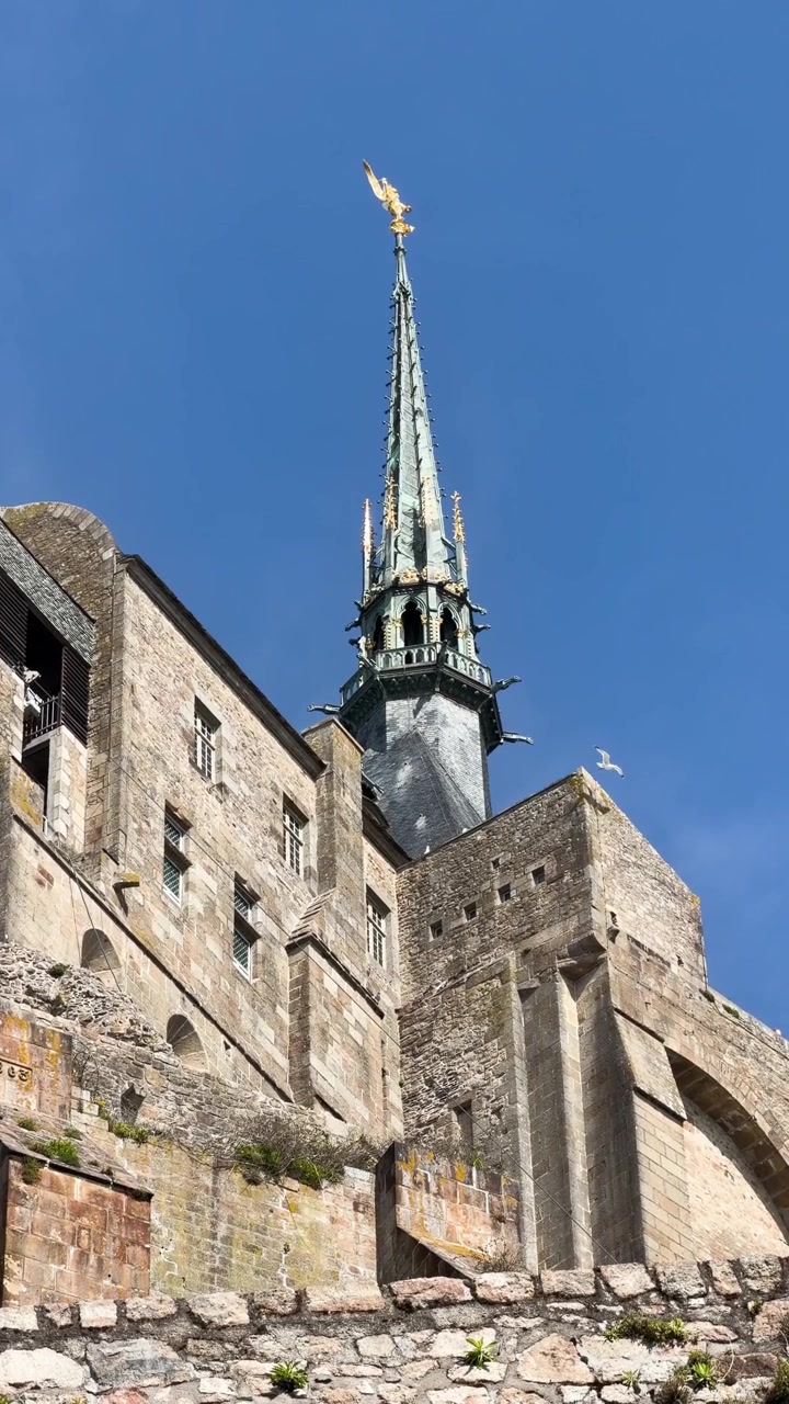 Opadvendt udsigt over Mont Saint-Michel klosterets spir og stenmure under klar blå himmel