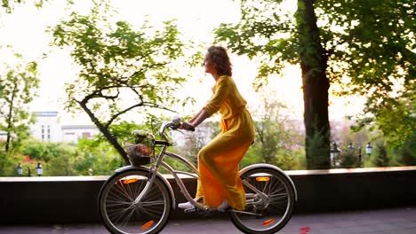 Brunette-smiling-woman-in-long-yellow-dress-is-enjoying-her-time-riding-a-city-bicycle-with-a-basket-and-flowers-inside-during