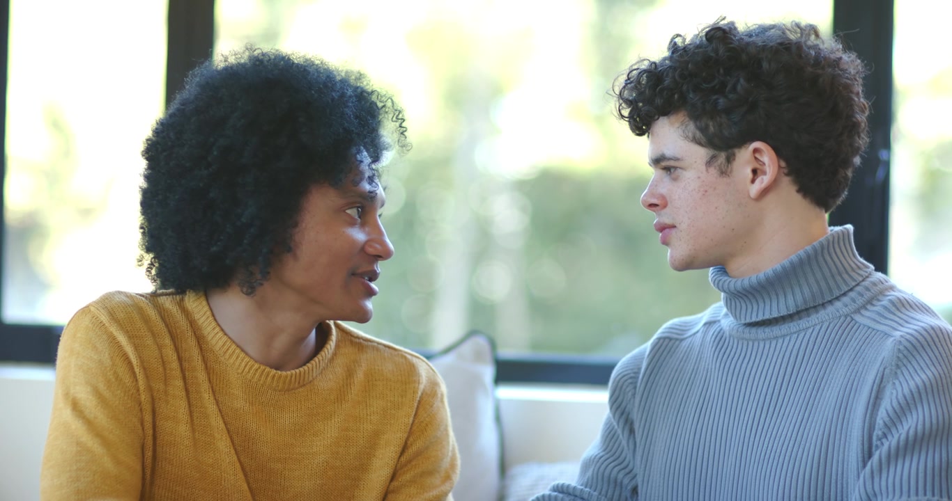 Diverse male friends sitting on sofa by window, mustard sweater leaning forward