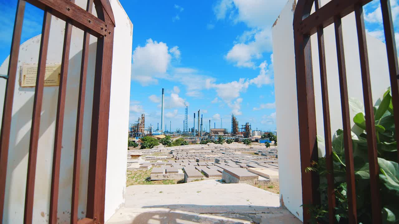 grandes puertas de hierro que conducen a un cementerio vacío en curacao con una gran refinería industrial al fondo