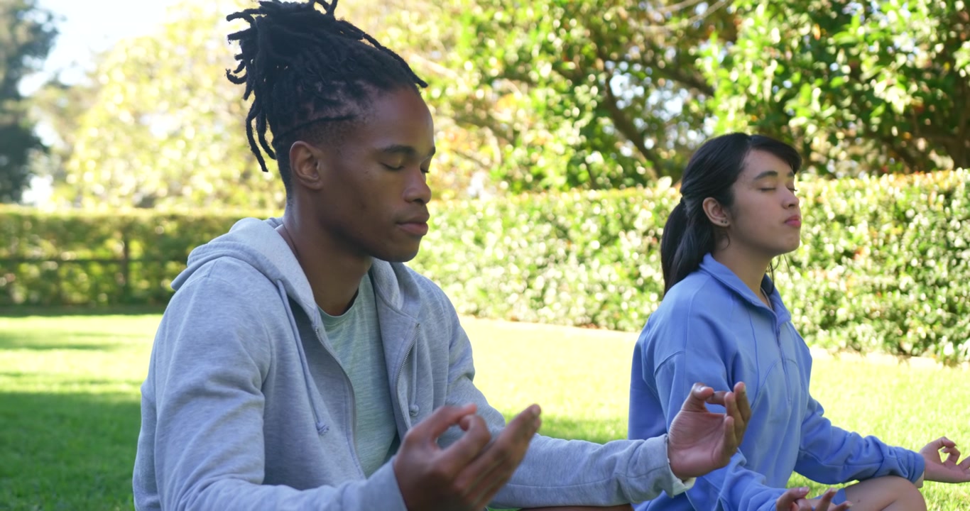 African American man and Asian woman meditating on grassy lawn in park, closing eyes, forming mudra