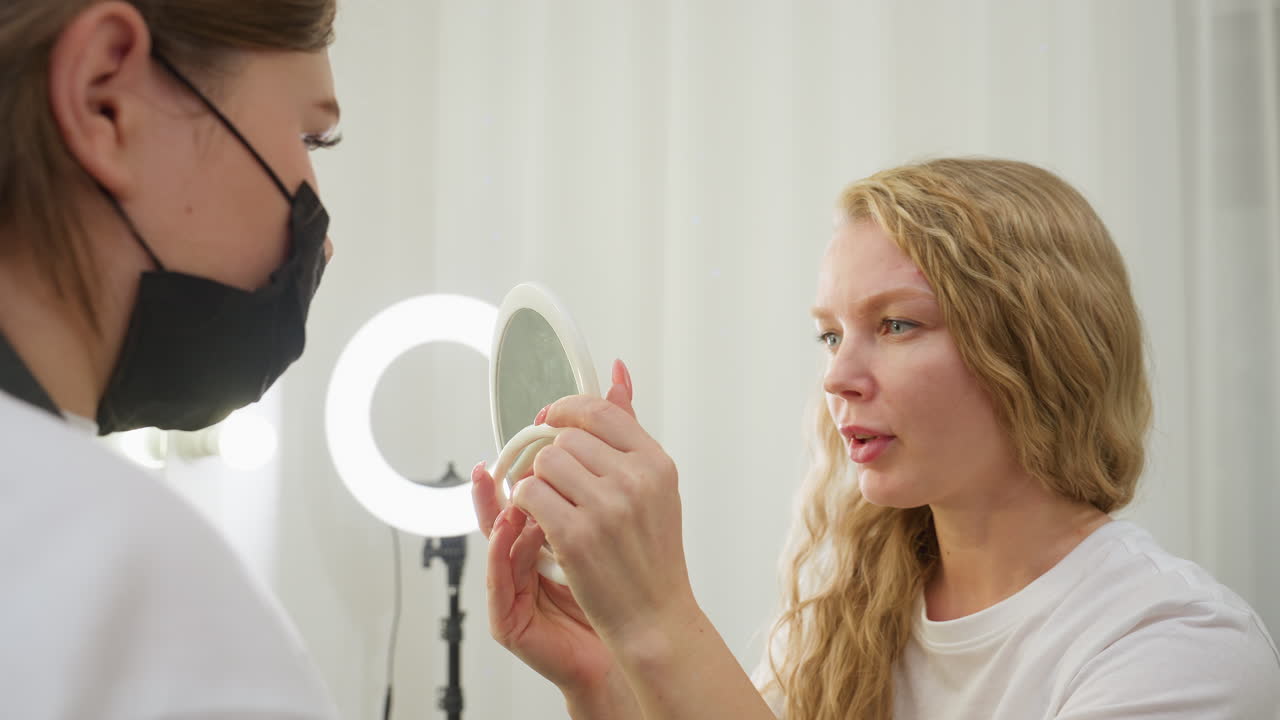 Blonde client carefully examines her freshly lined eyebrow using handheld mirror while seated in softly lit beauty studio. Focused expression reflects attention to eyebrow detail and cosmetic results