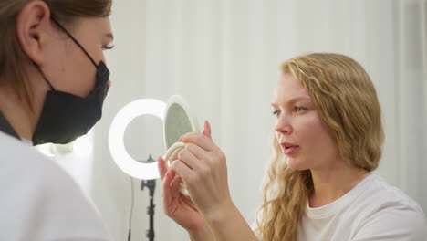 blonde client carefully examines her freshly lined eyebrow using handheld mirror while seated in softly lit beauty studio. focused expression reflects attention to eyebrow detail and cosmetic results