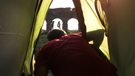 young man backpacker exits camping tent in front of roman aqueduct arches in parco degli acquedotti park ruins in rome at sunset sticks in ground shot from inside tent camera slow motion pov