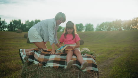 mother seated with her young daughter on a hay bale, sharing a moment together as the girl holds a brown cover book, surrounded by a tranquil field