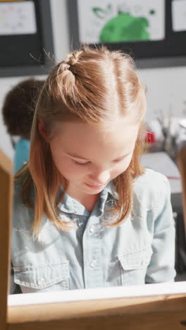 vertical video of happy caucasian schoolgirl sitting at easel painting in diverse class, copy space