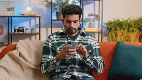 Indian-hindu-young-man-sitting-on-sofa,-using-smartphone-share-messages-on-social-media-application