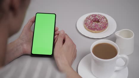 woman sitting at table coffee donut using smartphone with chroma key green screen, scrolling through social network media online shop internet. smartphone in horizontal mode with green screen mock-up.