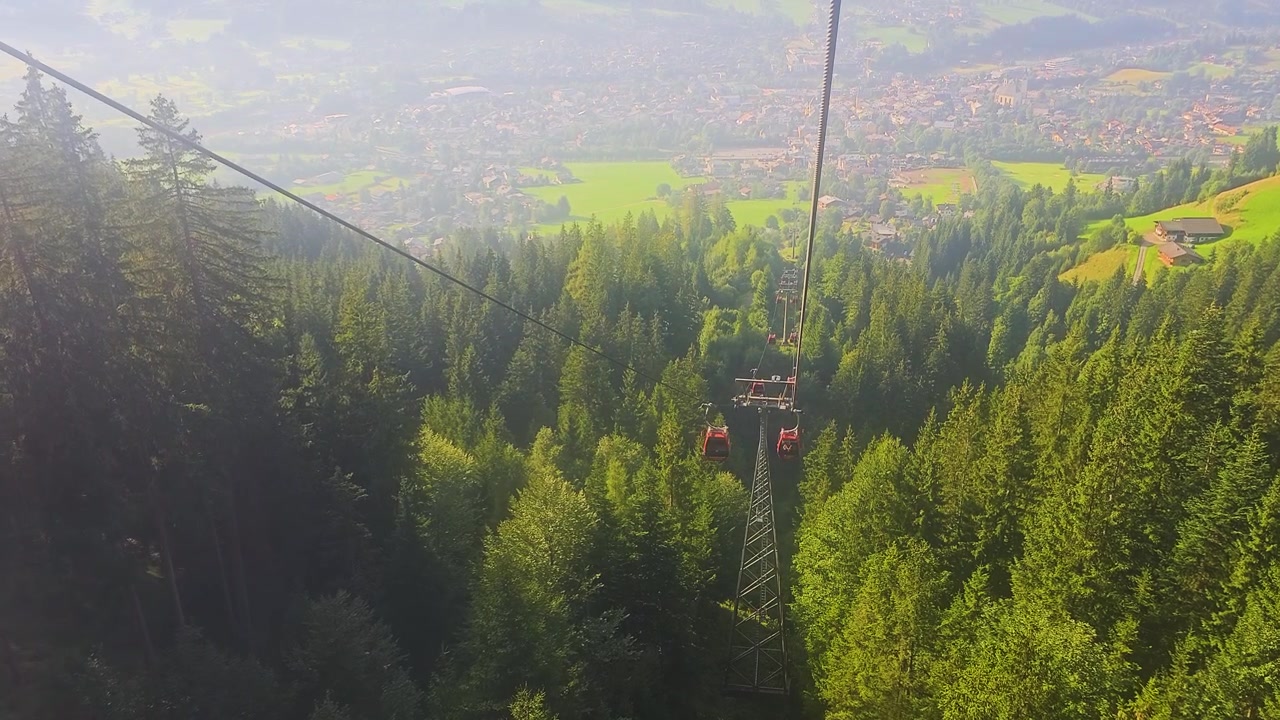 Veduta di viaggio da una gondola che si affaccia su una città di montagna, hotel alpini, prati verdi, una foresta di conifere e catene montuose distanti durante una soleggiata vacanza estiva in una scena di turismo europeo.