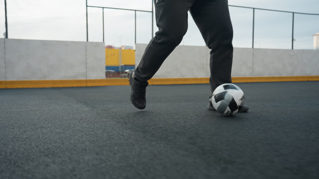 Leg view of man skillfully training with soccer ball on sport arena during a dynamic practice session, background includes urban buildings, fencing, and goalpost