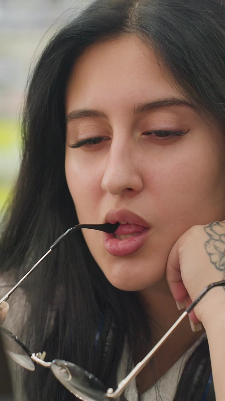 Primo piano di una giovane donna con lunghi capelli scuri e occhiali che si aggiusta. La donna ha tatuaggi sul polso e interagisce da vicino con un oggetto metallico rotondo. La vegetazione sfocata suggerisce un parco.