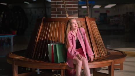 lady wearing pink outfit drops her shopping bags near a wooden sit out in a mall, she holds a black handbag sits down with her left hand resting on the bench, legs slightly crossed