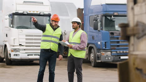 distant view of a boss and worker wearing vests and safety helmets organizing a truck fleet in a logistics park while they consulting a document 1