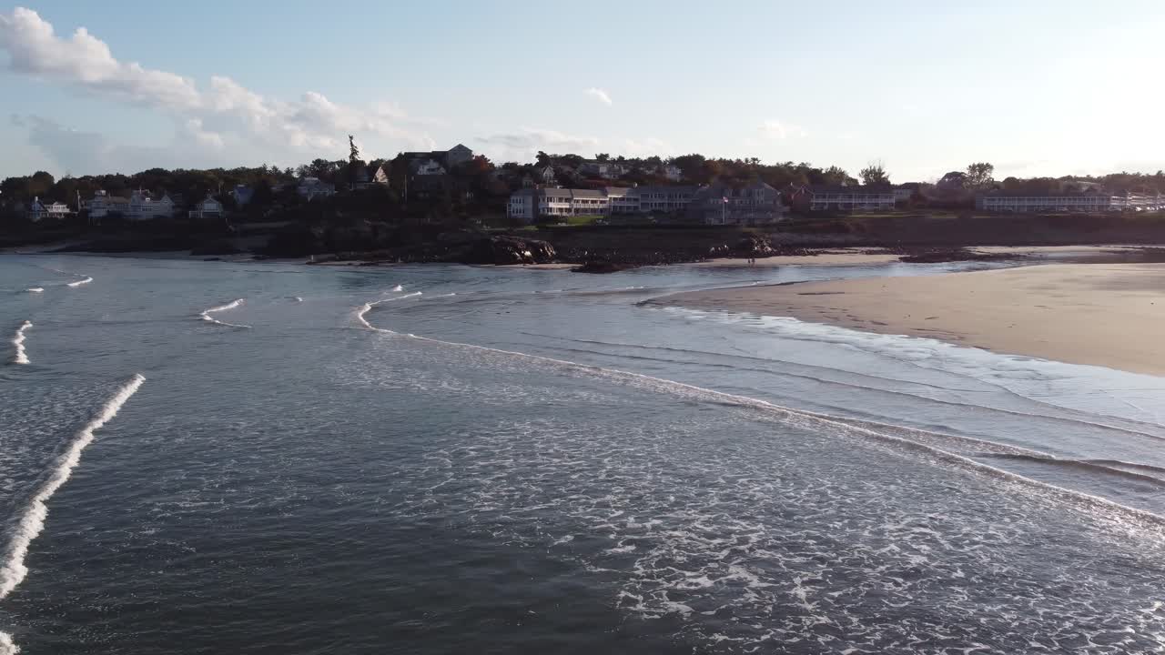 Drone che vola sopra l'acqua dell'oceano con lo skyline di Ogunquit in lontananza durante il tramonto, vista aerea della costa nel Maine, USA.