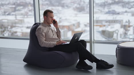 young businessman sits in bean bag chair, talks on the phone and surfing his laptop. panoramic windows with blurred winter cityscape at backdrop.