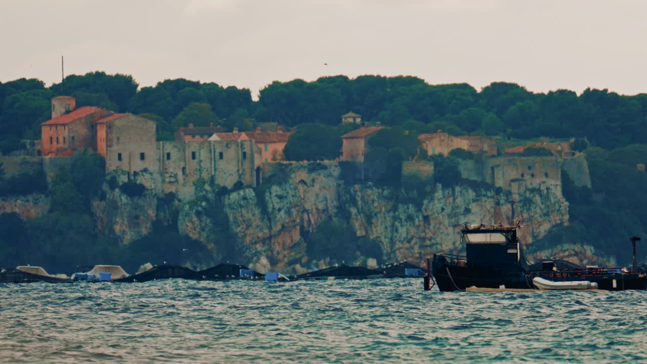 A medieval stone fortress and village perched on a high cliff above the Mediterranean Sea, with a fish farm and moored platforms spread across the water below