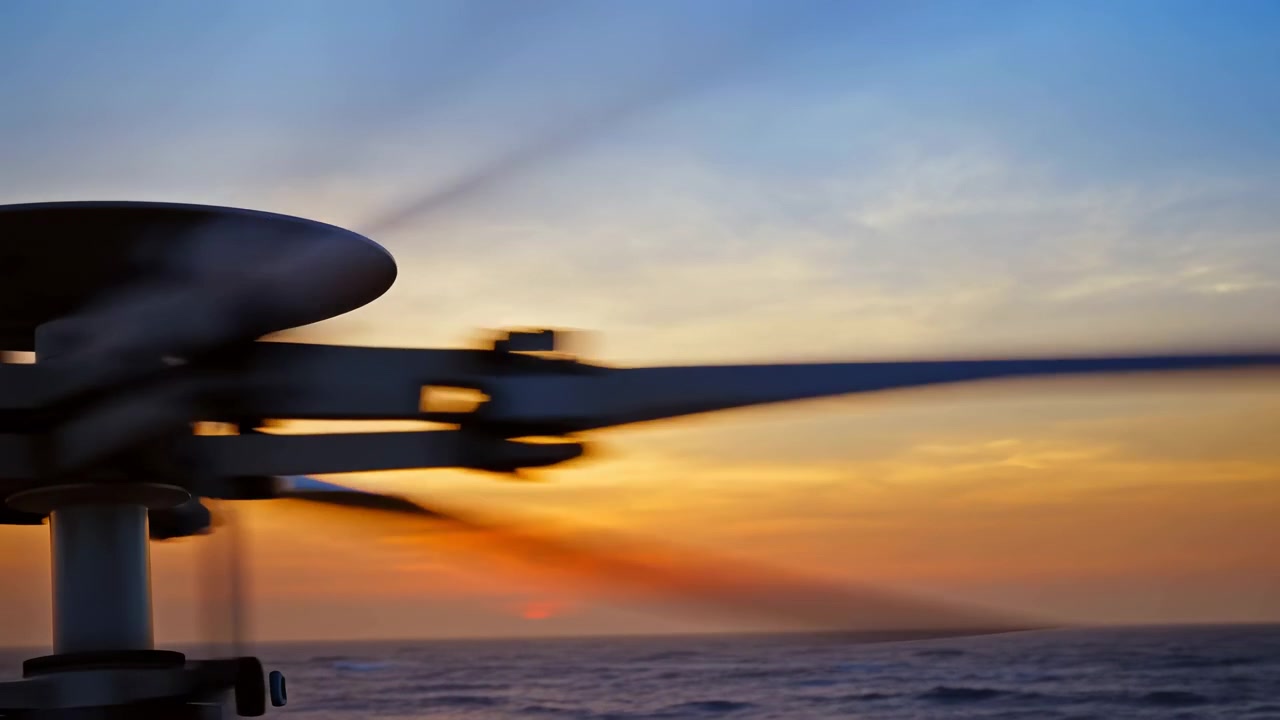 Slørede rotorblade fra en drone ses i venstre forgrund som en mørk silhuet, med varmt solnedgangslys, bløde skyer, en klar horisont og roligt vand.
