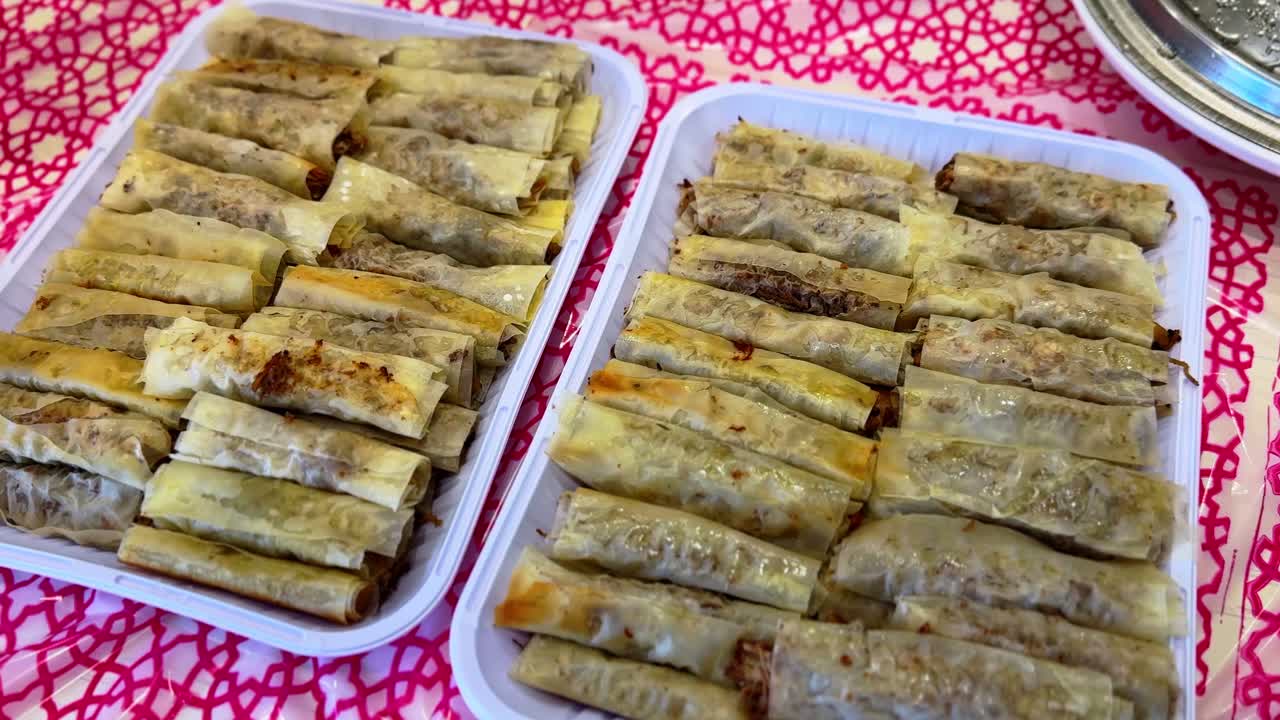 Primo piano di involtini primavera di carne croccanti appena preparati, disposti ordinatamente in vassoi di plastica su una tovaglia rossa a motivi, pronti per essere serviti a un raduno o un pasto in famiglia.