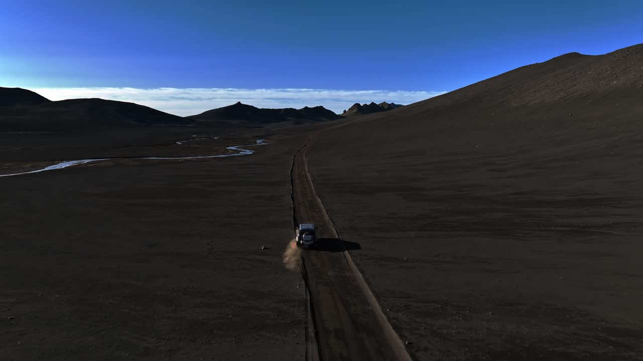 Il drone segue un 4x4 che sbanda leggermente fuori dalla pista sterrata attraverso una valle vulcanica nera, con un fiume serpeggiante in lontananza e montagne di fronte sotto un cielo blu.