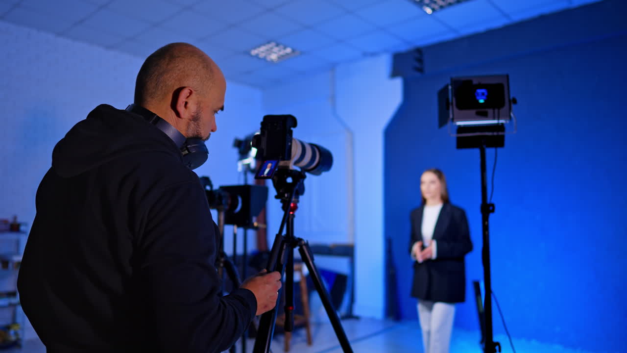 Over the shoulder view of videographer filming woman in white suit. Cameraman records female presenter standing against blue background with professional studio lights and monitor setup