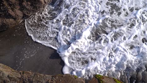 Top-down-view-of-sandy-beach-and-rocks-with-waves-crashing-in-slow-motion