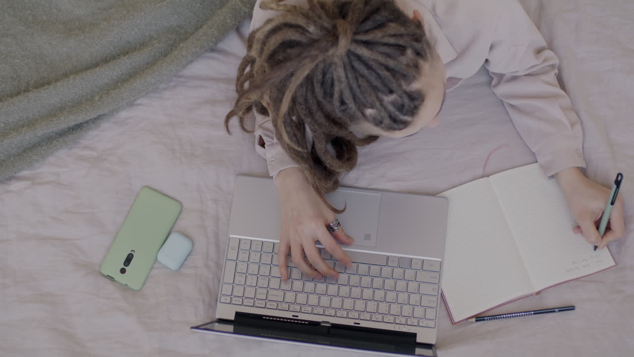 Woman working from bed with laptop, notebook, and phone