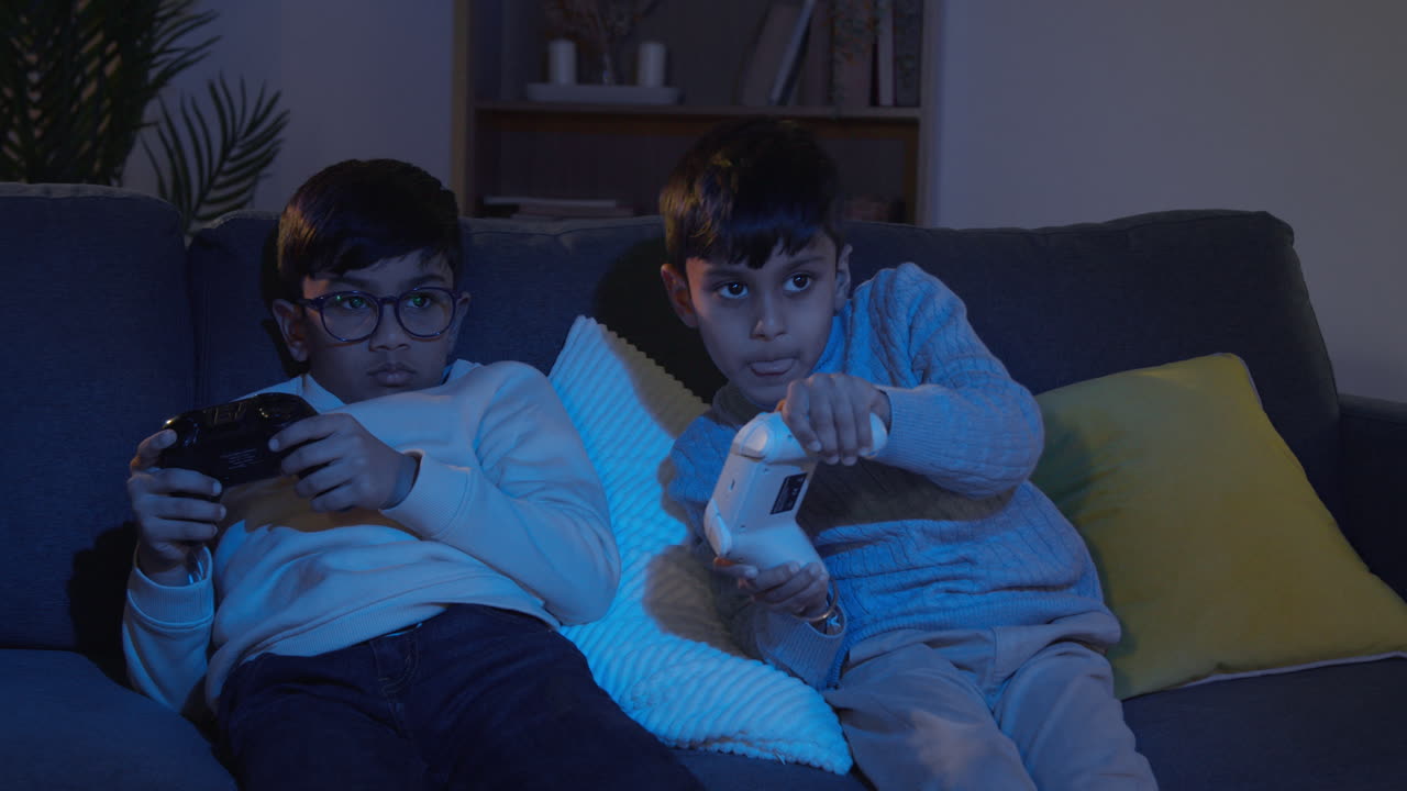 Two Young Boys Sitting On Sofa At Home Playing With Computer Games Console On TV Holding Controllers Late At Night