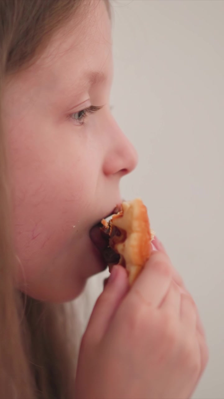 Close up of young girl eating food holding and bringing pastry to her mouth. scene shows detailed interaction with baked goods against plain background lighting