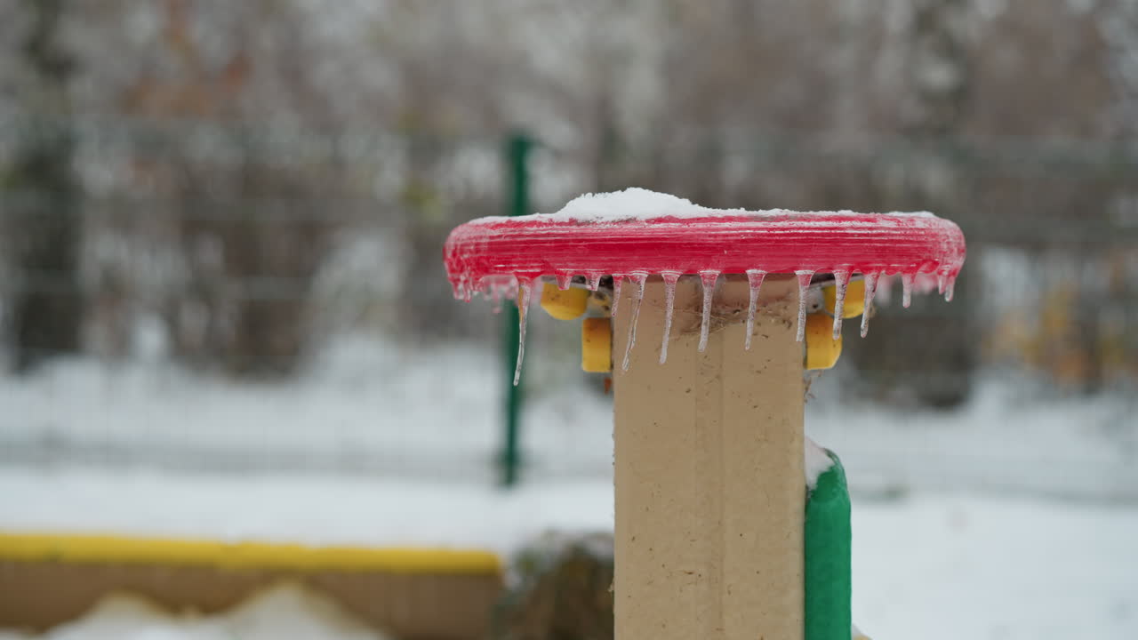 놀이터 구조에 매달린 얼음 조각과 함께 은 원형 얼음 표면의 클로즈업, 얼음 세부 사항의 대조와 흐릿한 눈 바탕을 포착, 멀리 자동차의 흐릿한 시야